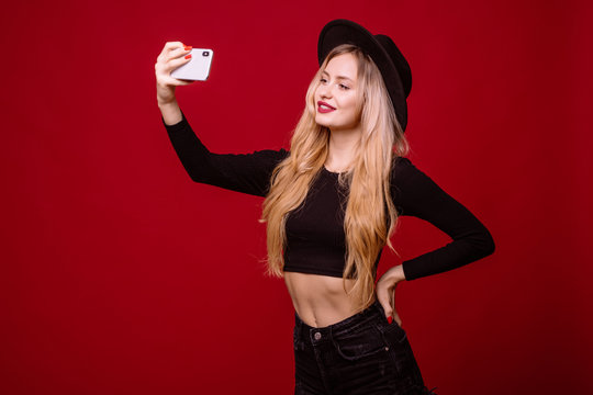 Cool blonde woman in black t-shirt and hat taking selfie on her cell phone isolated over red background
