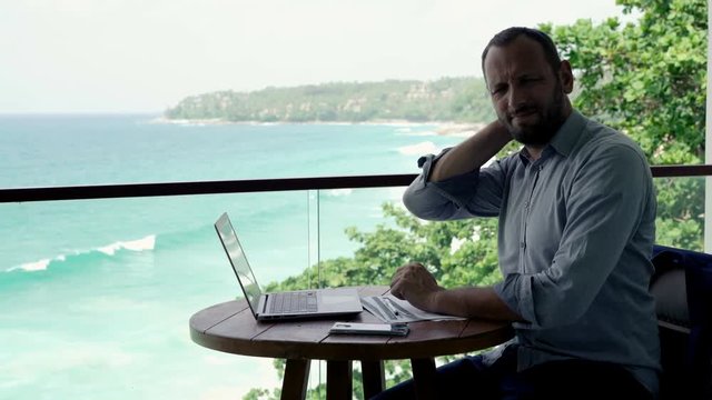 Young businessman having neck pain during work on laptop on terrace, with sea view, 4K
