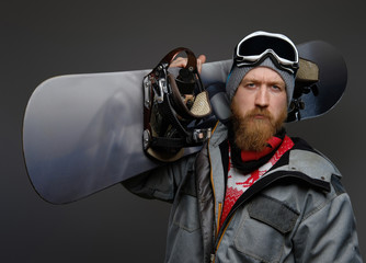 Confident man with a red beard wearing a full equipment holding a snowboard on his shoulder, looking at a camera with a serious look, isolated on a dark background.