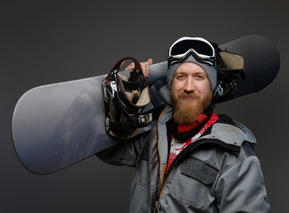 Confident man with a red beard wearing a full equipment holding a snowboard on his shoulder, smiling and looking at a camera, isolated on a dark background.