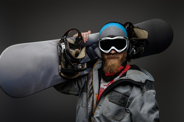 Brutal man with a red beard wearing a full equipment holding a snowboard on his shoulder, isolated on a dark background.