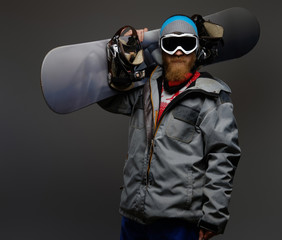 Brutal man with a red beard wearing a full equipment holding a snowboard on his shoulder, isolated on a dark background.