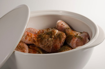 Raw chicken drumsticks marinated in spices in a large white ceramic saucepan with a lid