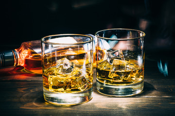 Two glasses of scotch whiskey or cognac with ice cubes and bottle of alcohol liquor on dark wooden background