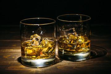 Two glasses of scotch whiskey or cognac and ice cubes on dark wooden background