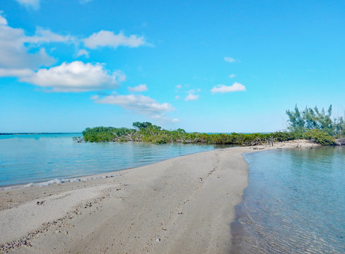 A pebbled sandbar breaking the surf of aquamarine seas with lush tropical scrub bushes spreading across the end under bright blue skies with puffy white clouds. Taken in the Eleutheras of the Bahamas.