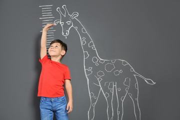 Cute little child measuring height near chalk giraffe drawing on grey background