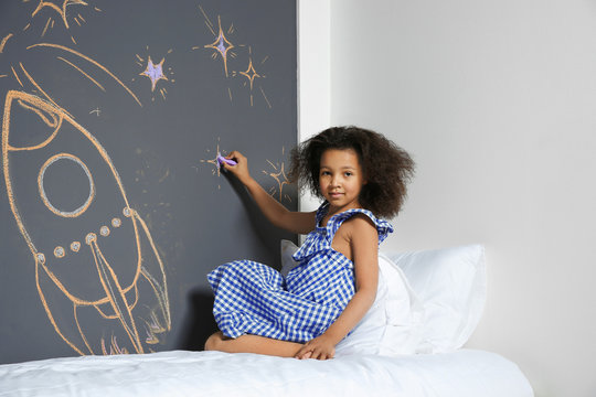 African-American child drawing rocket with chalk on wall in bedroom