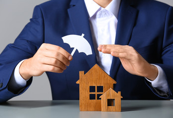 Man covering wooden houses with umbrella cutout at table, closeup. Home insurance