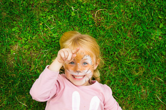 beautiful little girl at the children's party with a butterfly pattern on her face, posing emotionally while lying on the bright green grass