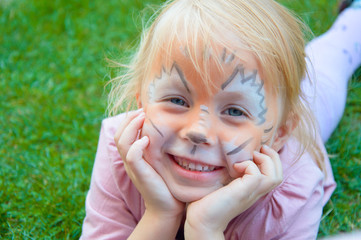 beautiful little girl blonde on a children's holiday with a pattern of a lion cub on her face, emotionally posing while lying on bright green grass holding a flower in her teeth