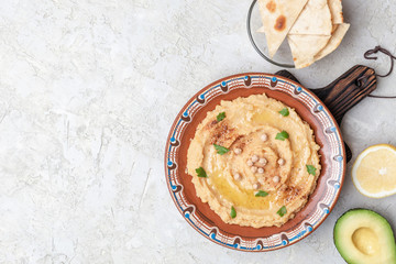 Hummus in to a brown clay plate with a blue pattern. On the white table are vegetables, greens, triangular pieces of pita. Top view. Flat lay.
