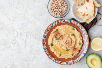 Hummus in to a brown clay plate with a blue pattern. On the white table are vegetables, greens, triangular pieces of pita. Top view. Flat lay.