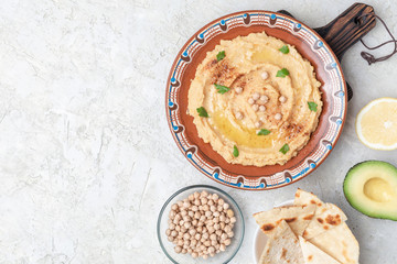 Hummus in to a brown clay plate with a blue pattern. On the white table are vegetables, greens, triangular pieces of pita. Top view. Flat lay.