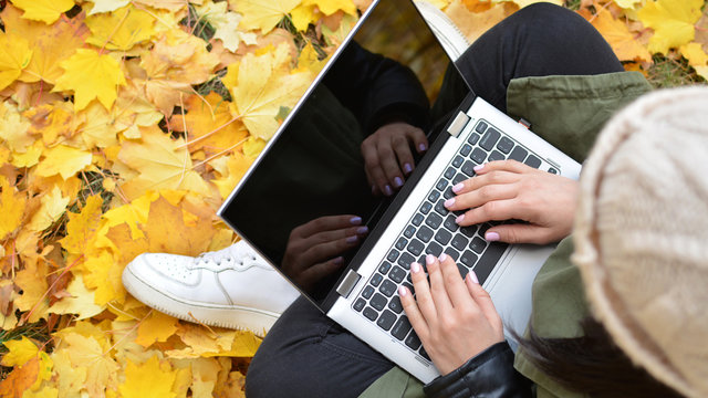 Girl in hipster with laptop in autumn park. A woman in a cap using a laptop while sitting on fallen leaves. Freelancer in the hat uses remote communication technology. Remote work. View from above