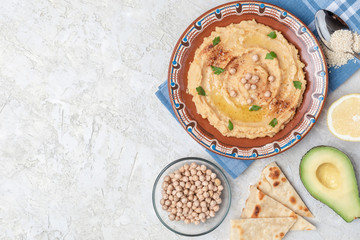 Hummus in to a brown clay plate with a blue pattern. On the white table are vegetables, greens, triangular pieces of pita. Top view. Flat lay.