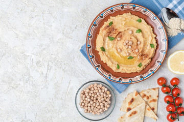 Hummus in to a brown clay plate with a blue pattern. On the white table are vegetables, greens, triangular pieces of pita. Top view. Flat lay.