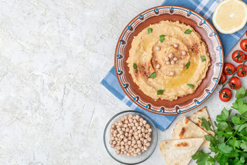 Hummus in to a brown clay plate with a blue pattern. On the white table are vegetables, greens, triangular pieces of pita. Top view. Flat lay.