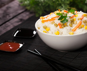 Cooked white rice with vegetables in a bowl with sauces and chopsticks on the wood black bamboo background.
