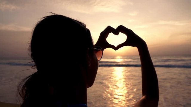 Woman making heart symbol with her hands during sunset on beach, super slow motion
