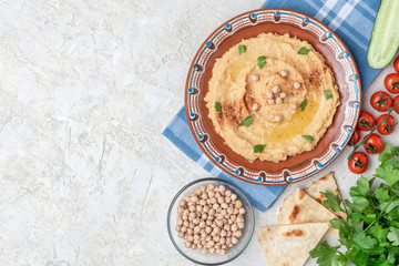Hummus in to a brown clay plate with a blue pattern. On the white table are vegetables, greens, triangular pieces of pita. Top view. Flat lay.
