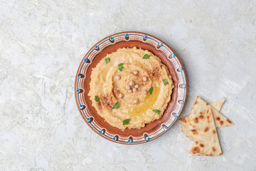 Hummus in to a brown clay plate with a blue pattern. On the white table are vegetables, greens, triangular pieces of pita. Top view. Flat lay.