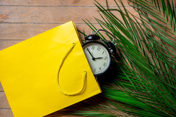 Fototapeta premium Vintage alarm clock in yellow bag and palm leaves on wooden table. Above view