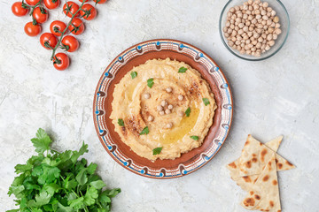 Hummus in to a brown clay plate with a blue pattern. On the white table are vegetables, greens, triangular pieces of pita. Top view. Flat lay.