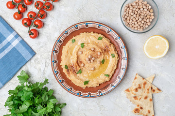 Hummus in to a brown clay plate with a blue pattern. On the white table are vegetables, greens, triangular pieces of pita. Top view. Flat lay.