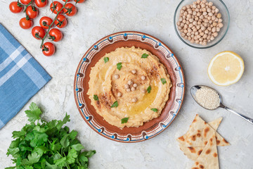 Hummus in to a brown clay plate with a blue pattern. On the white table are vegetables, greens, triangular pieces of pita. Top view. Flat lay.