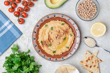 Hummus in to a brown clay plate with a blue pattern. On the white table are vegetables, greens, triangular pieces of pita. Top view. Flat lay.