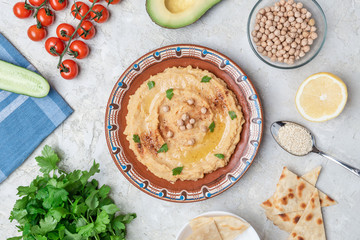 Hummus in to a brown clay plate with a blue pattern. On the white table are vegetables, greens, triangular pieces of pita. Top view. Flat lay.