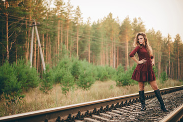Beautiful dreamy girl with curly natural hair enjoy nature in forest on railway. Dreamer lady in burgundy dress walk on railroad. Inspired girl on rails at dawn. Sun in hair in autumn. Good mood.