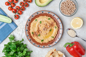 Hummus in to a brown clay plate with a blue pattern. On the white table are vegetables, greens, triangular pieces of pita. Top view. Flat lay.