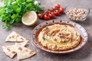 Hummus in a brown clay plate with a blue pattern. On the brown table are vegetables, greens, triangular pieces of pita. 