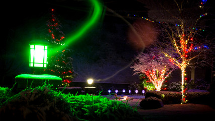 Holiday Photo of Lights in a Snow Covered Neighborhood at Night - with Green and Yellow Streaks Shooting from the Street Lamps and Multiple Trees Lined with Traditional, Multi Colored Lights