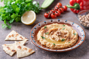 Hummus in a brown clay plate with a blue pattern. On the brown table are vegetables, greens, triangular pieces of pita. 