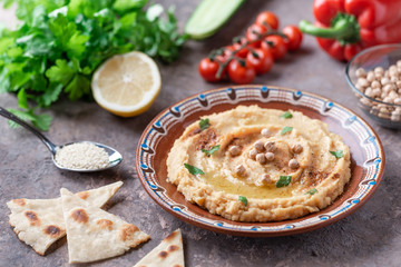 Hummus in a brown clay plate with a blue pattern. On the brown table are vegetables, greens, triangular pieces of pita. 