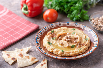 Hummus in a brown clay plate with a blue pattern. On the brown table are vegetables, greens, triangular pieces of pita. 