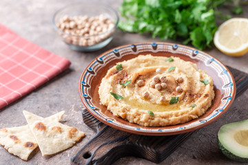 Hummus in a brown clay plate with a blue pattern. On the brown table are vegetables, greens, triangular pieces of pita. 
