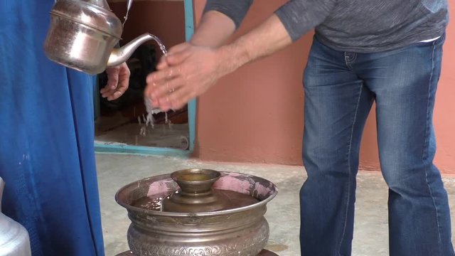 Inside his house Moroccan berber man wearing traditional muslim clothes is pouring water on European Caucasian man's hands