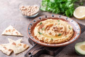 Hummus in a brown clay plate with a blue pattern. On the brown table are vegetables, greens, triangular pieces of pita. 