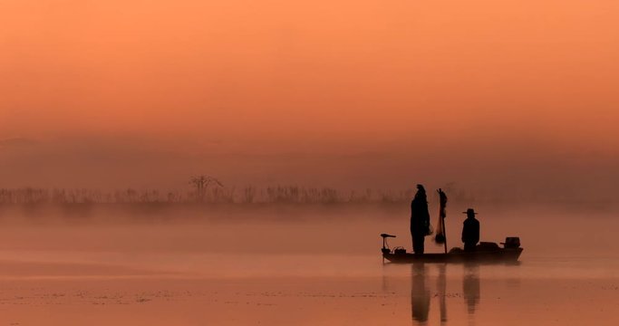 Traditional asian fishermen in silhouette using nets to fish at an exotic river location from a flat-bottomed boat at sunrise or sunset