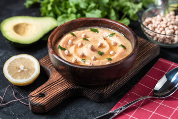 Hummus in to a brown wooden plate. On the black stone table are vegetables, greens, triangular pieces of pita. Top view. 