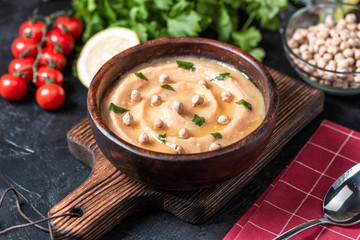 Hummus in to a brown wooden plate. On the black stone table are vegetables, greens, triangular pieces of pita. Top view. 