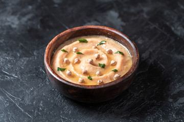Hummus in to a brown wooden plate. On the black stone table are vegetables, greens, triangular pieces of pita. Top view. 