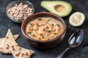 Hummus in to a brown wooden plate. On the black stone table are vegetables, greens, triangular pieces of pita. Top view. 