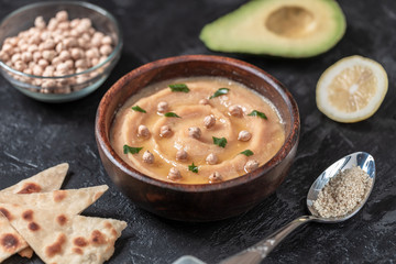 Hummus in to a brown wooden plate. On the black stone table are vegetables, greens, triangular pieces of pita. Top view. 