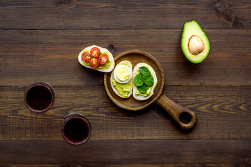 Fototapeta na wymiar Light appetizer with avocado. Toasts with vegetables and guacamole near glasses of wine on dark wooden background top view