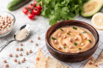 Hummus in the brown wooden plate. On the white table are vegetables, greens, triangular pieces of pita.